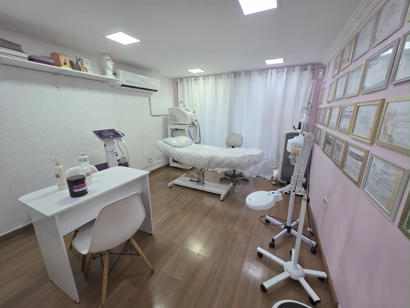 Sala de atendimento da clínica: maca branca, aparelhos, cadeira e mesa brancas, cortina clara e uma parede rosa coberta de certificados emoldurados.